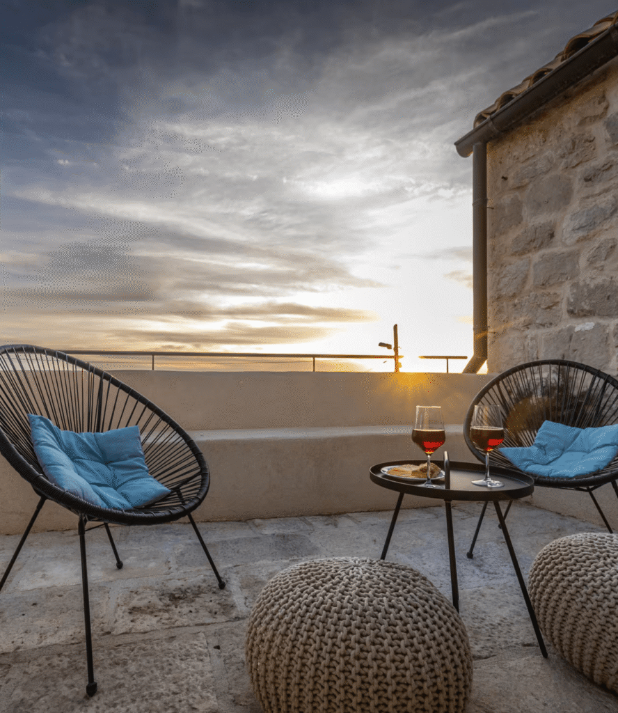 Outdoor rooftop terrace at sunset featuring black wicker chairs with blue cushions, a small round table with two glasses of red wine and snacks, and a stone wall in the background.