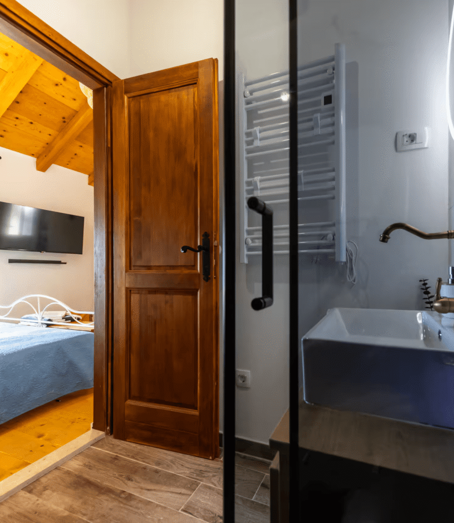 Open wooden door from a cozy bedroom into a bathroom with a white sink and a wall-mounted towel warmer visible behind a glass partition.