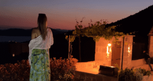 Woman looking out over the Dubrovnik coastline at dusk from Villa Brsečine's terrace, with candlelit stone archways and a vine-covered pergola beside her.