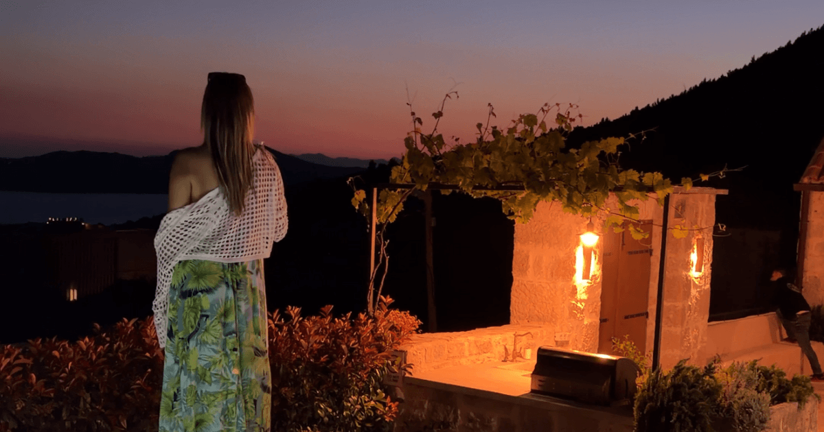 Woman looking out over the Dubrovnik coastline at dusk from Villa Brsečine's terrace, with candlelit stone archways and a vine-covered pergola beside her.